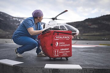 Une professionnelle manipule un conteneur de transport médical rouge contenant une greffe humaine urgente, avec un hélicoptère en arrière-plan sur une piste d'atterrissage.