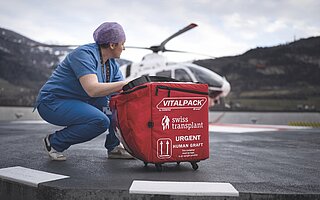 Une professionnelle manipule un conteneur de transport médical rouge contenant une greffe humaine urgente, avec un hélicoptère en arrière-plan sur une piste d'atterrissage.